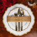 A Posh Setting white plastic plate with gold scalloped bands, silverware, and a napkin on a table.