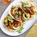 A white plate with a taco filled with avocado and corn on a tortilla with a plate of grilled vegetable and corn tacos.