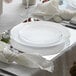 A white table setting with a Posh Setting silver mirrored charger plate, white plate, and silverware on a table.