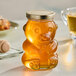 A glass bear jar of honey on a table next to a cup of tea.