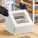 A person in black gloves putting a box of food into a white Baker's Lane box with a window.