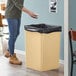 A woman standing in front of a Lavex beige square trash can with a black garbage bag inside.