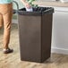A woman wearing tan pants puts a black plastic bag in a brown Lavex square trash can.
