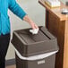 A brown Lavex square trash can with a drop shot lid, shown in use with a person disposing of a tissue.