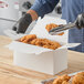A white take-out box with a handle being filled with pieces of fried chicken using tongs.