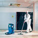A person in a white protective suit and mask using a Nilfisk wet/dry vacuum to clean a room.