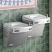 A stainless steel bi-level drinking fountain with two spouts mounted on a tiled wall.