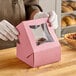 A person in gloves opening a pink Baker's Lane box with a clear window to reveal a pastry.