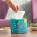 A hand putting a tissue in a blue Kleenex Naturals box with white dots on a table.