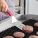 A hand holding the Choice Perforated Turner with a red handle cooking burgers on a grill.