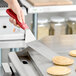 A hand using a Choice square edge turner with a red handle to cut a pancake on a griddle.