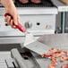 A hand using a Choice square edge turner with a red handle to lift bacon from a grill.