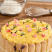 A cake decorated with yellow pastry cream, pink flower petals, and herbs, surrounded by ladyfinger biscuits.