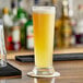 A glass of yellow liquid on a yellow surface with a glass of beer on a coaster.
