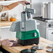 A commercial continuous feed food processor with a green and gray base, shown in use on a kitchen counter with food being processed into a container.