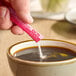 A hand holding a pink and white ecoStick pouring powder into a cup of coffee.