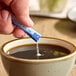 A hand holding an ecoStick Blue packet of powder over a cup of coffee.