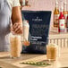 A 3.5 lb. bag of Capora Pumpkin Latte Frappe Mix displayed on a counter with prepared pumpkin latte drinks.