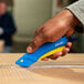 A hand holding a Pacific Handy Cutter blue and yellow safety knife to cut cardboard.