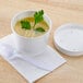 A white Huhtamaki poly paper food cup filled with soup and a plastic lid with a spoon on the table.