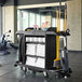 A Rubbermaid black and white housekeeping cart with cleaning supplies including towels and a broom.