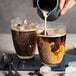 A person pouring milk into a Duralex glass of brown liquid.