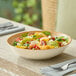 A round beige porcelain deep coupe plate filled with a colorful salad, placed on a dining table.