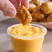 A hand holding a small piece of pretzel dipping into a cup of Gehl's Aged Cheddar Cheese Sauce.