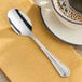 An Acopa Edgeworth stainless steel teaspoon on a saucer next to a cup of coffee.
