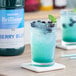 A glass of Monin Brilliance Berry Blue natural energy drink with blue liquid and berries on a table.