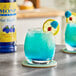 A glass of blue raspberry-flavored drink garnished with a lemon slice, a blue candy, and a red berry, with a bottle of Monin Sugar-Free Blue Raspberry Flavoring Syrup in the background.