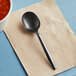 A black Choice medium weight polypropylene plastic soup spoon on a beige napkin next to a bowl of soup.