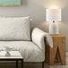 A 15-inch ceramic table lamp with a light concrete finish and a white linen shade, placed on a wooden side table next to a white sofa.