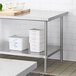 An adjustable stainless steel undershelf designed for 30-inch by 72-inch work tables, shown installed beneath a work table and holding food storage containers.