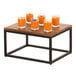 A Cal-Mil black metal rectangular display riser holding a glass of orange juice on a table with a purple flower in it.