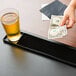 A person's hand holding a dollar bill over a black bar mat with a glass of beer on it.