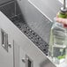 A hand using a Choice black plastic interlocking bar mat strip to hold a green bottle over a stainless steel sink.