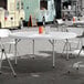 A round, white plastic bi-folding table with gray legs set up outdoors with folding chairs around it.