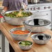 A set of ten heavy weight stainless steel mixing bowls in various sizes, some filled with salad ingredients, displayed on a wooden countertop in a kitchen setting.