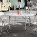 A round, white plastic bi-folding table with gray legs, set outdoors with folding chairs around it.