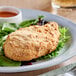 A white plate topped with a piece of breaded chicken on a table with a salad and a glass of liquid.