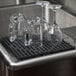 A group of glasses and cups on a black Choice interlocking bar mat on a counter over a sink.