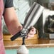 A person using a Kayser stainless steel whipped cream dispenser to add whipped cream to a drink.