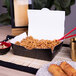 A box of Southern Champion black take-out containers filled with noodles on a table with a bowl of food and red chopsticks.