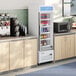 A white swing glass door merchandiser refrigerator with multiple shelves, stocked with beverages, situated between countertops in a break room or kitchen area.