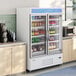 A white swing glass door merchandiser refrigerator filled with assorted beverages, situated between countertops in a commercial setting.