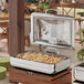 An outdoor catering table with an Acopa Manchester stainless steel chafer with a glass top and soft close lid full of food.