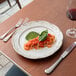A white round stoneware salad plate with a scalloped edge and embossed floral design, shown with a serving of pasta and a green garnish.