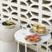 A round ivory melamine plate holding a sandwich, displayed on a white table next to a glass of water and another plate of food.