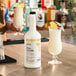 A 1-liter bottle of Hometown Provisions Piña Colada mix placed on a bar counter next to a prepared piña colada drink garnished with pineapple and a cocktail umbrella.